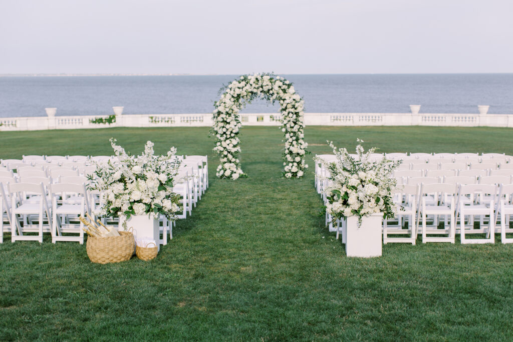 Outdoor ceremony setup overlooking the ocean, featuring a lush floral arch and white seating for guests, part of a full-service wedding weekend itinerary designed for a seamless and stunning celebration