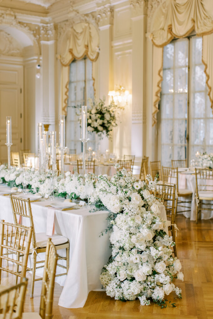 Elegant ballroom reception with gold chairs, white linens, cascading white floral arrangements, and tall candle centerpieces, designed as part of a full-service wedding weekend itinerary to create a cohesive, elevated guest experience