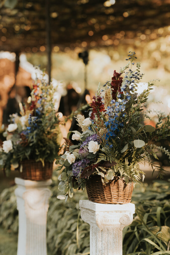 Wicker baskets filled with lush floral arrangements sit atop white columns in a garden ceremony setting.