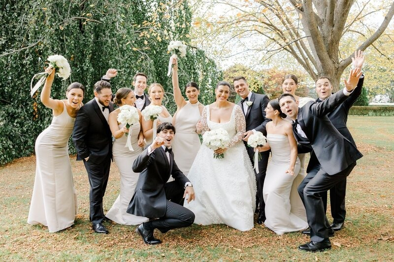 Joyful wedding party poses outdoors in formal attire, surrounded by autumn leaves and lush greenery.