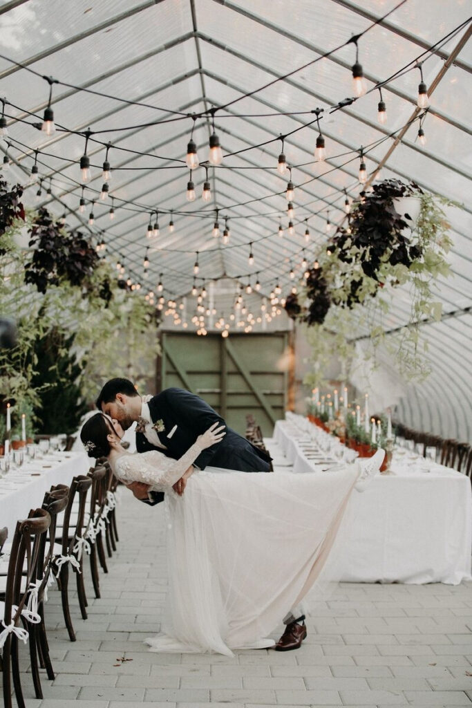 Romantic moment of a newlywed couple sharing a kiss under a glass tent filled with hanging greenery and string lights, captured during a wedding dinner setup—an example of full service wedding planning with curated design and seamless coordination.