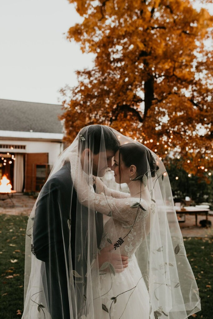 Bride and groom sharing an intimate moment under a veil in front of a cozy autumn backdrop with string lights and a fire pit—capturing the emotion and setting that full service wedding planning helps bring to life.