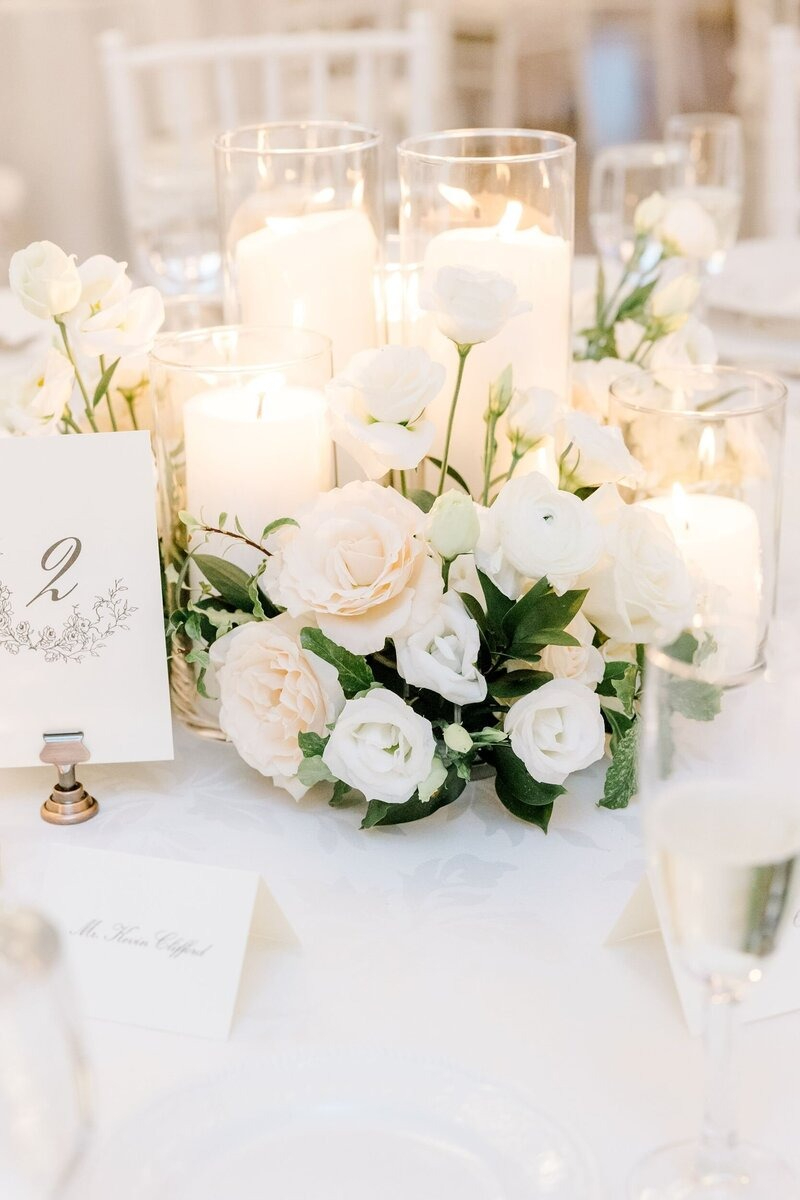 Romantic wedding table centerpiece with cream and blush roses, soft greenery, glowing pillar candles, and an elegant table number card in a calligraphy-style font.