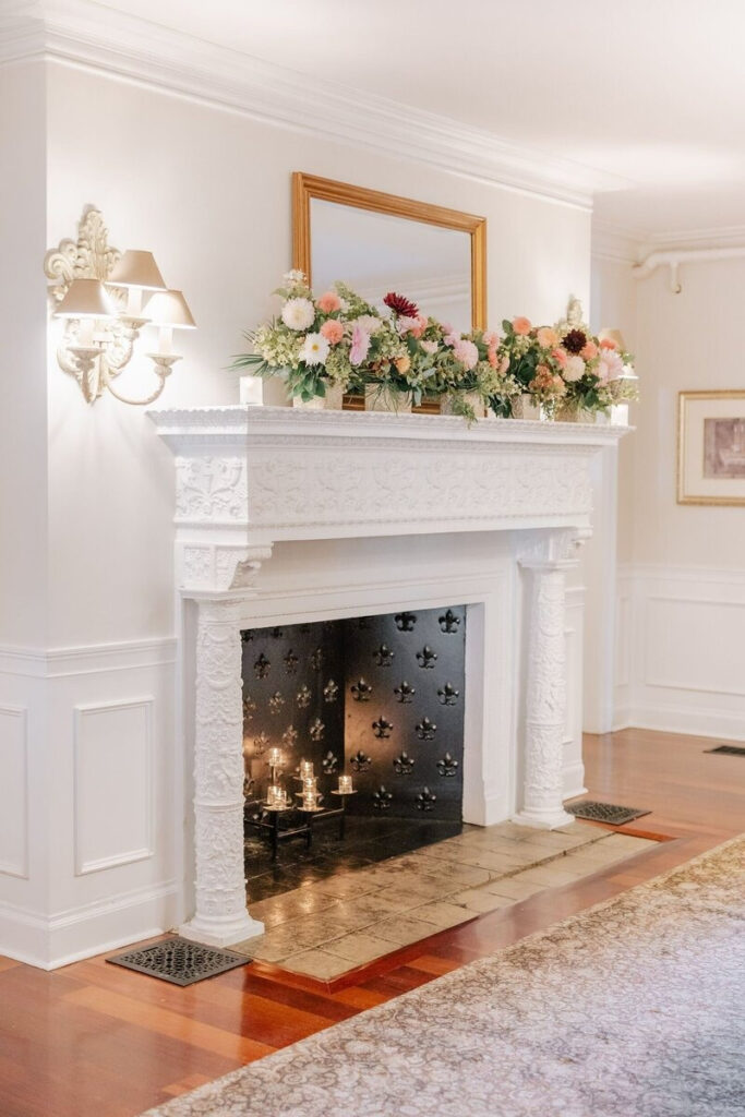 White carved fireplace decorated with soft pink and burgundy floral arrangements, glowing candlelight inside, and a gold-framed mirror above.