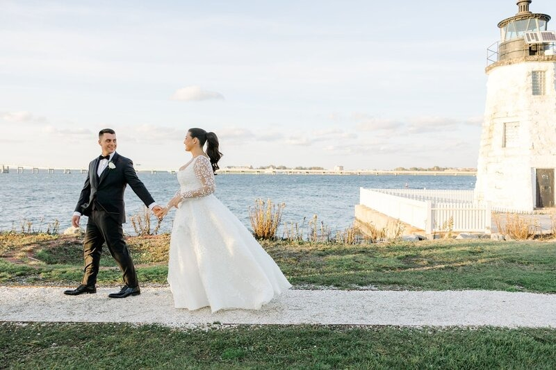 Bride and groom walking hand in hand by the water near a lighthouse during their New England wedding weekend, with soft sunset light behind them.