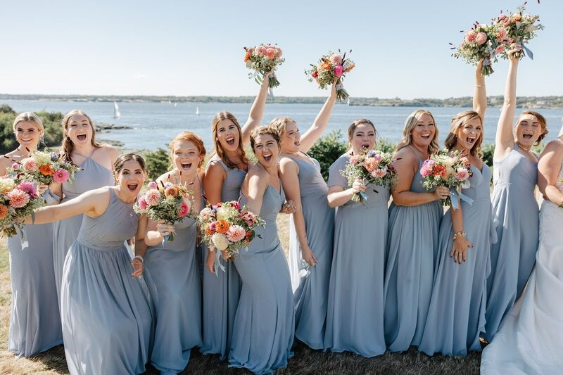 Joyful bridal party in soft blue dresses celebrating during a New England wedding weekend by the water, holding colorful floral bouquets.
