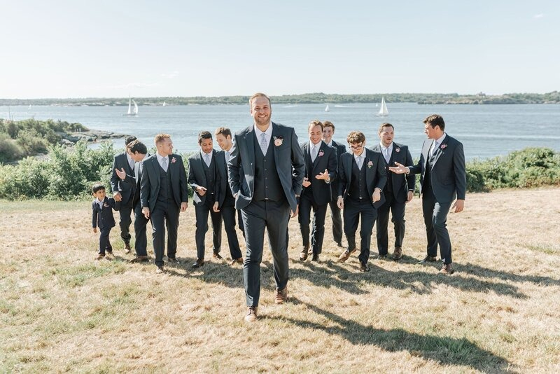 Groom and groomsmen in navy suits walking along the waterfront during a New England wedding weekend, with sailboats in the background.