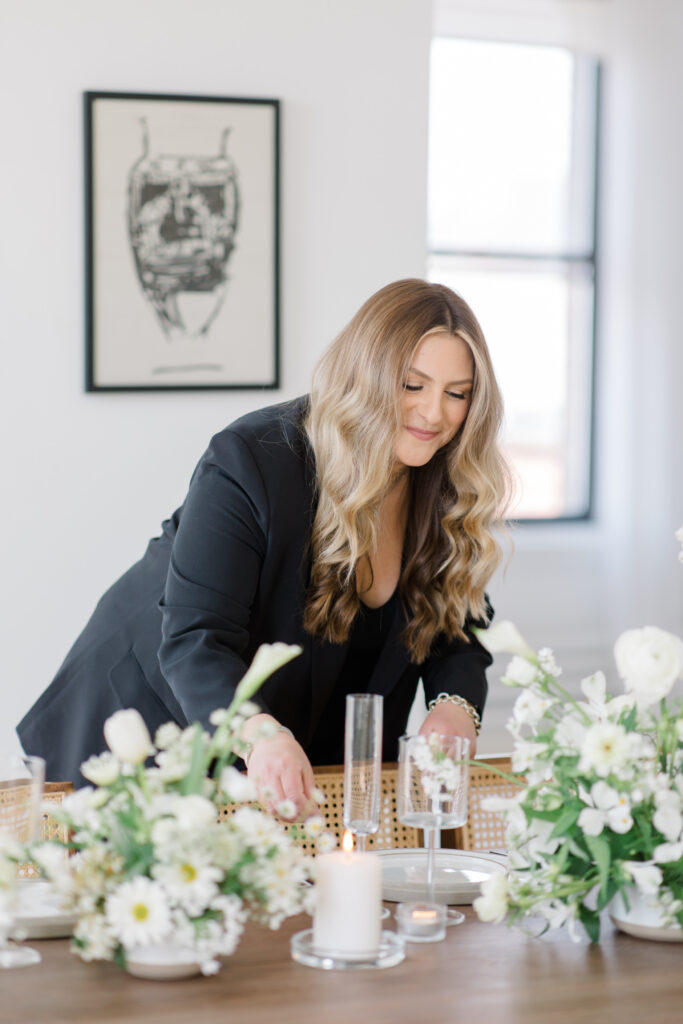 Wedding planner styling a table with white florals and candles, wearing a black blazer and smiling while adjusting decor in a bright, modern space.