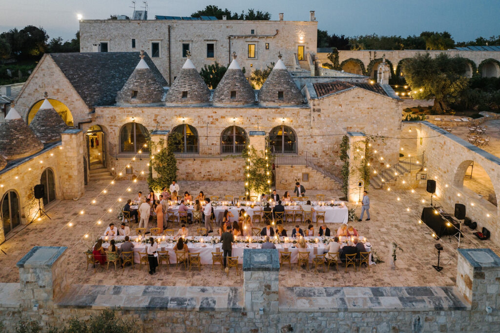 Evening courtyard wedding reception at a historic stone estate with trulli-style rooftops, long tables, and string lights glowing overhead.