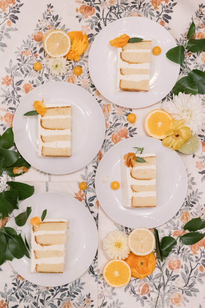Flat lay of white plates with slices of layered vanilla wedding cake, surrounded by citrus fruits, fresh greenery, and floral table linens.
