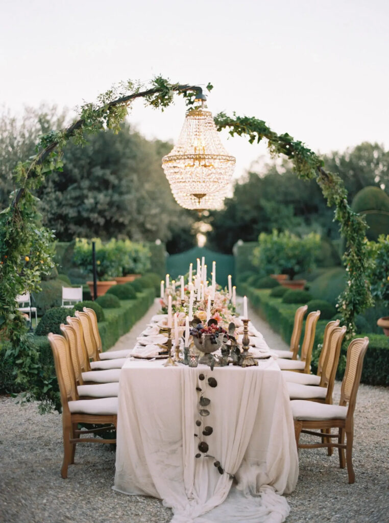 Elegant outdoor wedding reception table set at one of the villas in Italy, featuring neutral linens, vintage chairs, taper candles, and lush florals in a manicured garden setting.