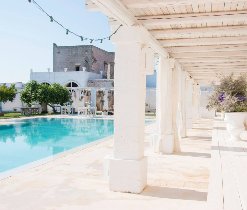 Bright and airy poolside terrace with white stone architecture, sheer curtains, and potted greenery at a traditional masseria in southern Italy.