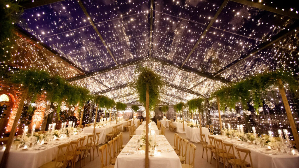 Romantic wedding reception setup at one of the villas in Italy, with long tables under a clear tent ceiling covered in twinkling string lights and hanging greenery.