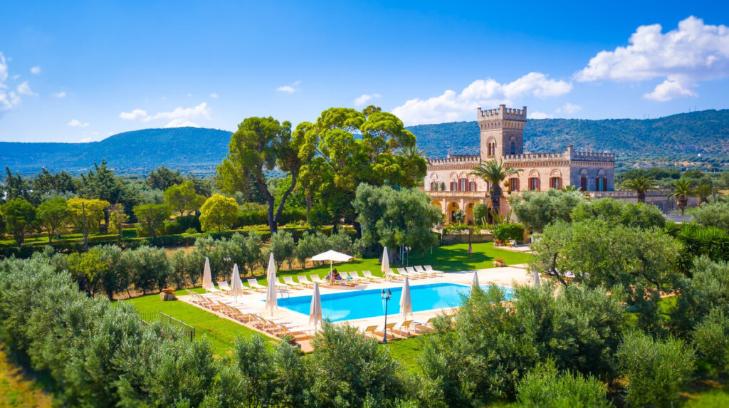 Lush estate with a castle-like villa, surrounded by olive trees and mountains, featuring a central pool area with lounge chairs and umbrellas.