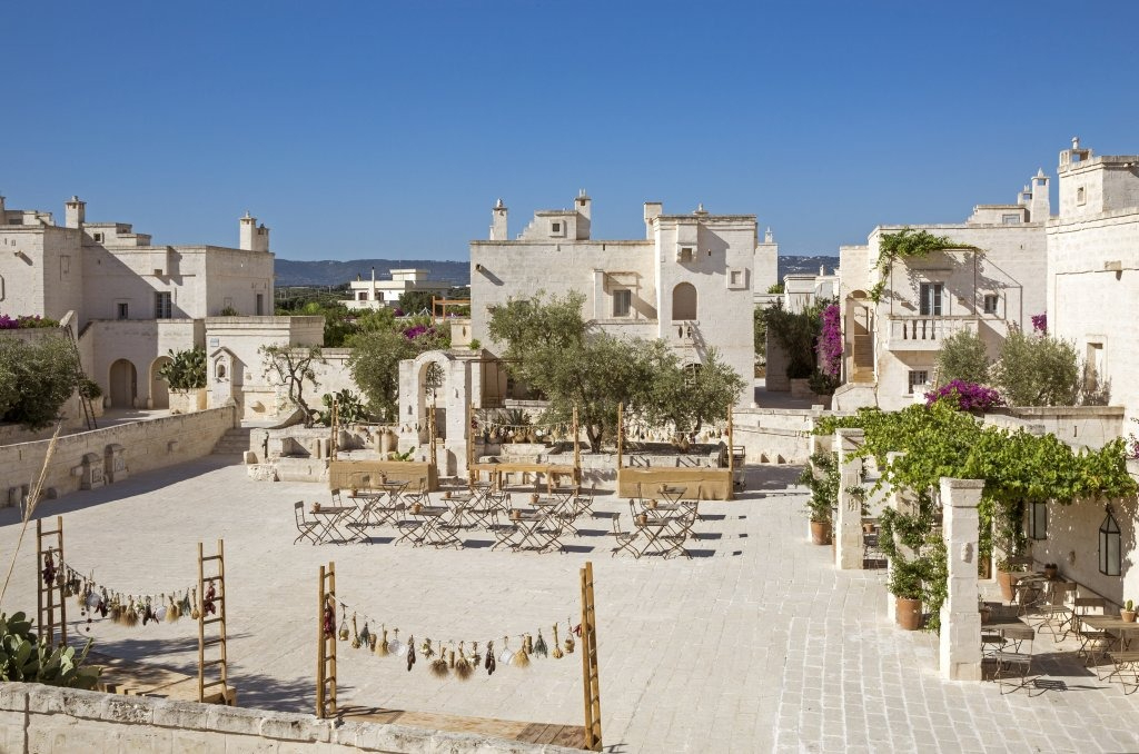 Open stone courtyard in a traditional southern Italian village, set up with wooden tables and chairs, olive trees, and string decor for an outdoor event.
