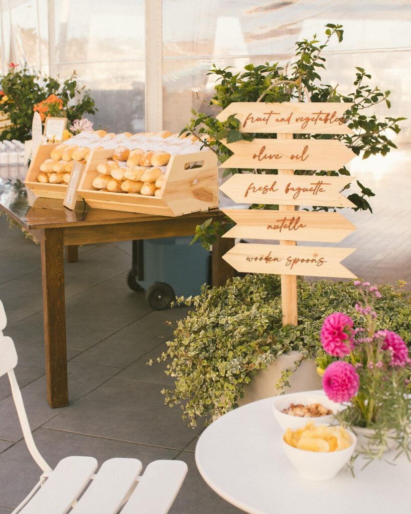 Rustic market-style wedding display with fresh baguettes, wooden directional sign, and vibrant floral accents.
