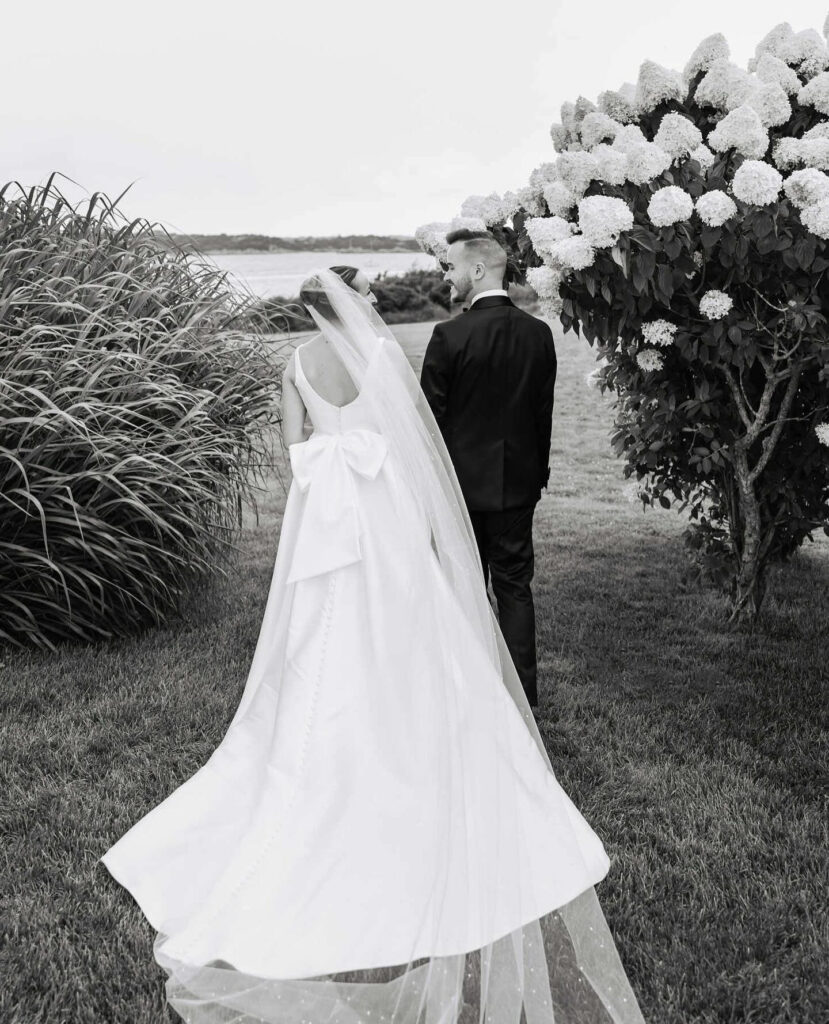 Black and white wedding photo of bride and groom walking outdoors beside blooming hydrangea bushes.