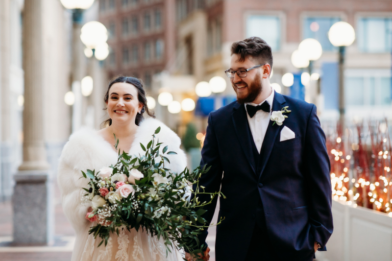 Bride in a white fur wrap and groom in a classic tux walk through city streets with glowing lights.
