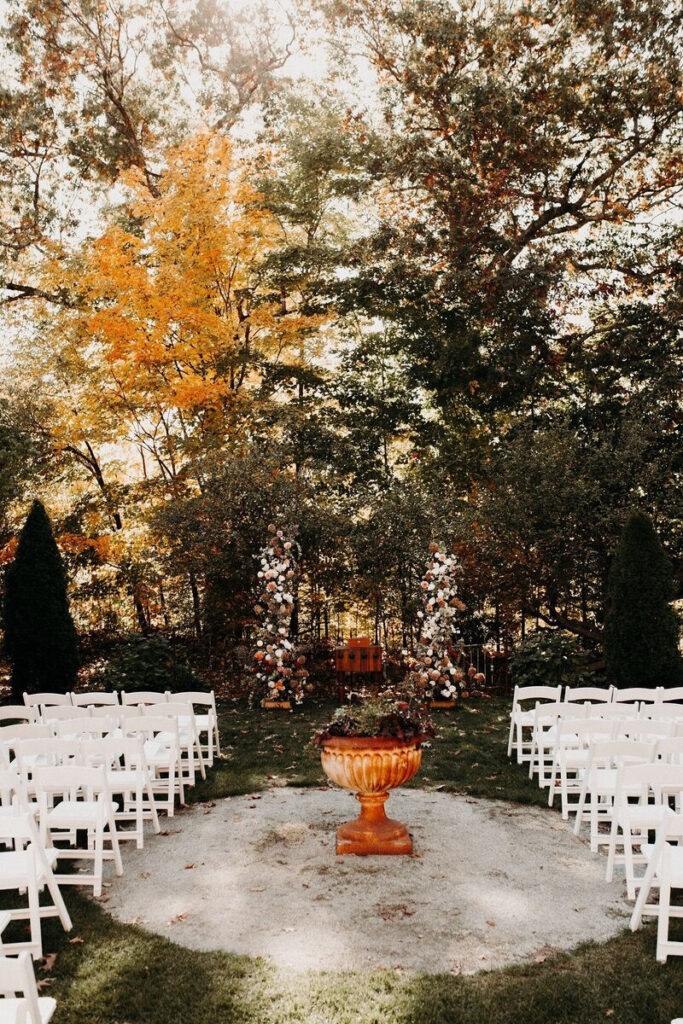 Outdoor fall wedding ceremony setup with white chairs, floral arrangements, and autumn trees as the backdrop.