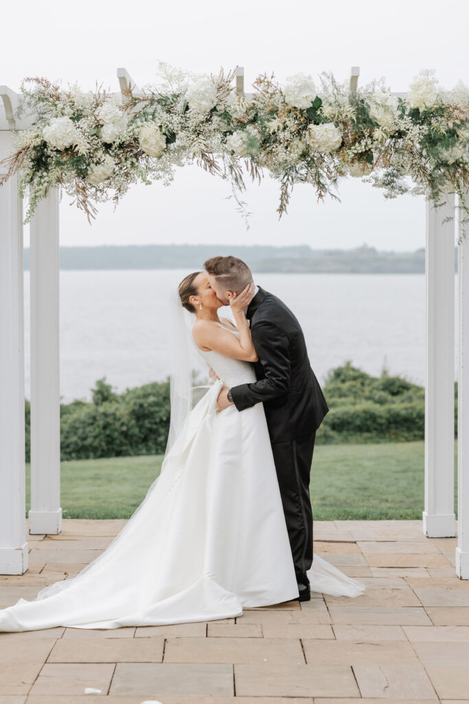 Bride and groom kissing under floral arch by the water, celebrating the best time of year to get married.