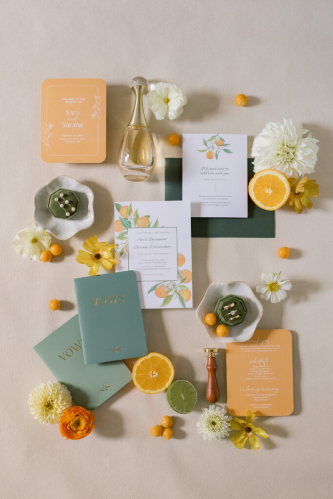 Flat lay of citrus-themed wedding stationery with vow books, invitations, flowers, rings, perfume, and sliced oranges and limes arranged on a light background.