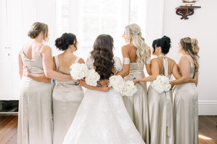 Bride with her bridesmaids holding white bouquets, all facing away in elegant light-toned dresses.
