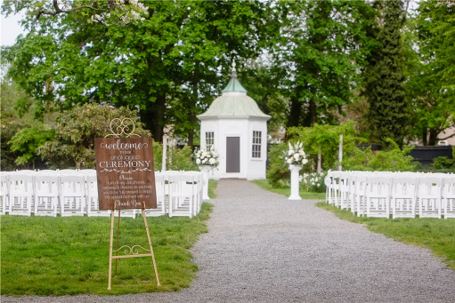 Outdoor garden ceremony setup with white chairs, floral arrangements, and a charming white pavilion at the end.