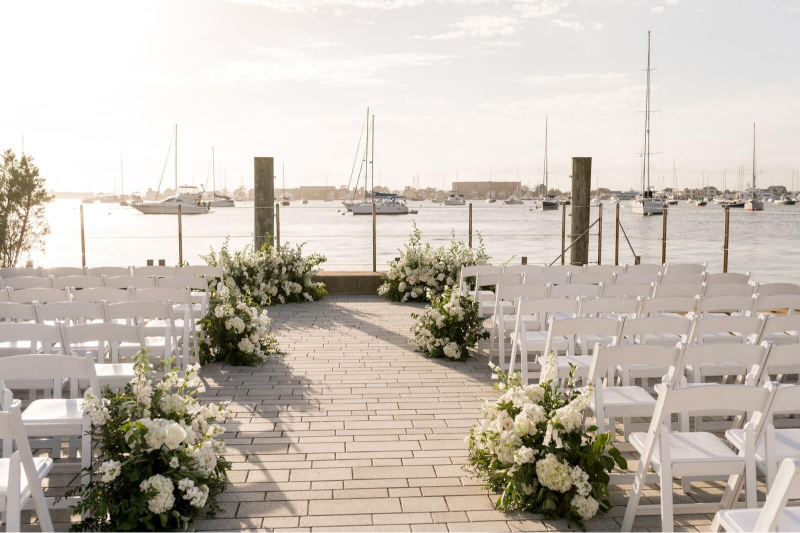 Waterfront ceremony setup with white chairs and floral arrangements overlooking a harbor filled with sailboats.
