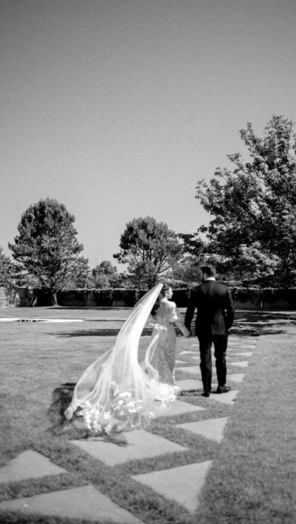 Black and white photo of a bride and groom walking hand in hand across a garden path.