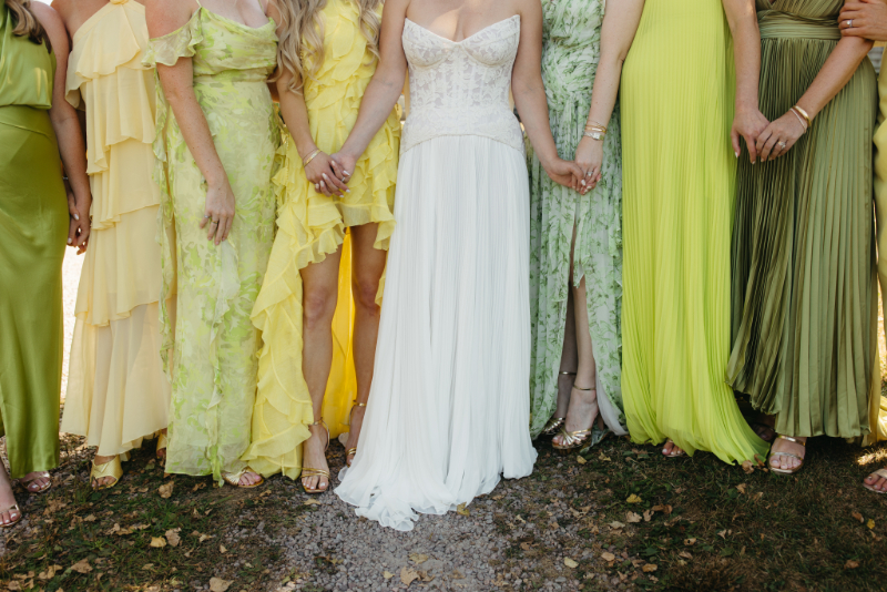 Bridal party in mismatched green and yellow dresses standing hand-in-hand with the bride in a flowing gown.