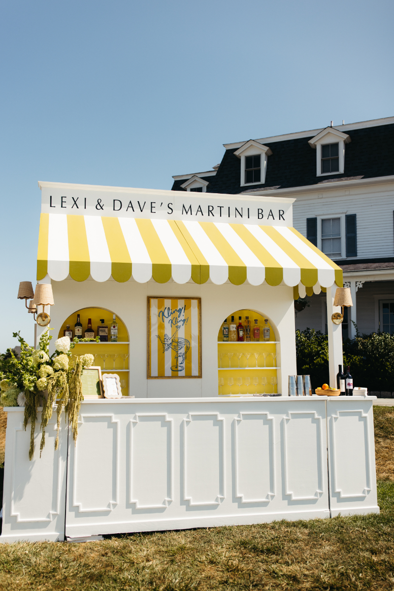 Charming outdoor martini bar with yellow and white striped canopy, custom signage, and playful floral accents.