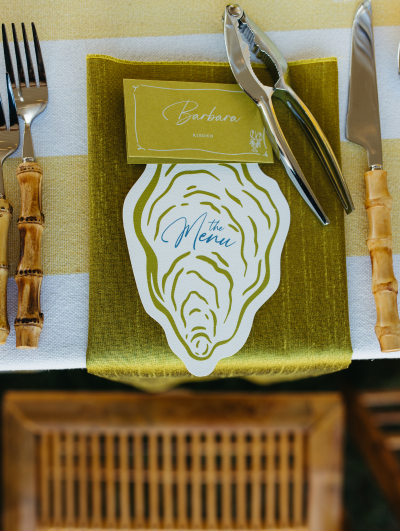 Oyster-shaped menu atop green textured linen with bamboo flatware and striped tablecloth for a coastal-inspired tablescape.