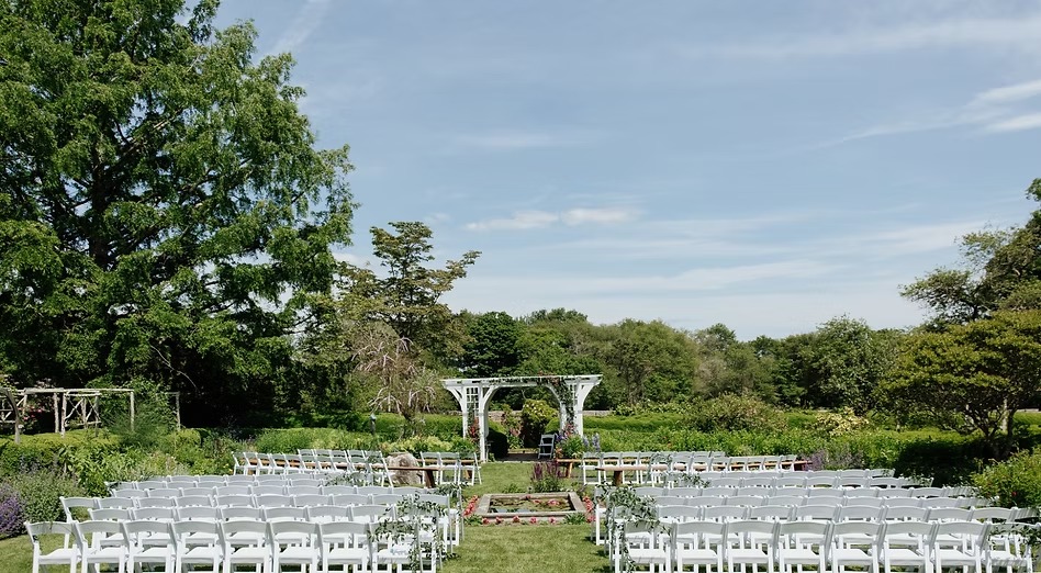 Outdoor garden ceremony setup with white chairs facing a white arbor, surrounded by lush greenery and blue sky.
