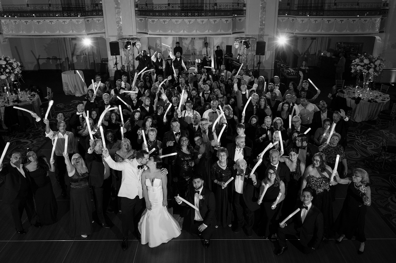 Black and white photo of a joyful wedding crowd on the dance floor holding glow sticks in celebration.