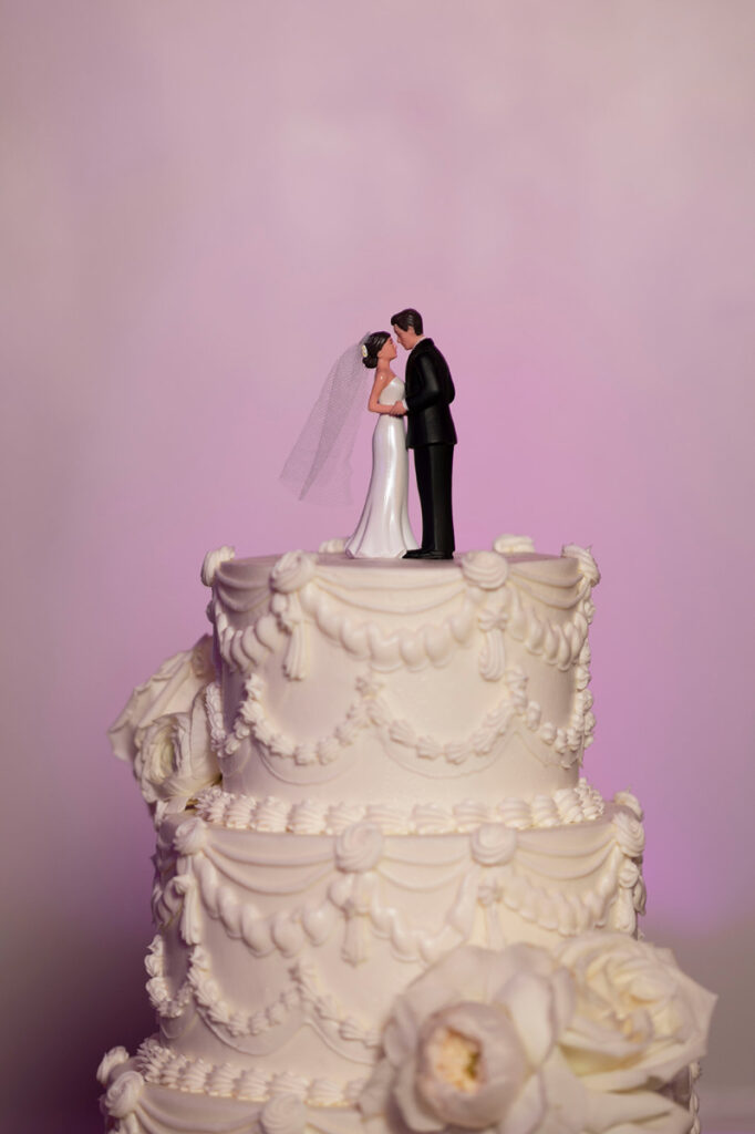 White tiered wedding cake with piped frosting details and a classic bride and groom topper against pink lighting.