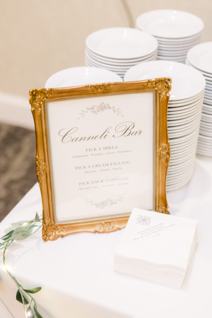 Cannoli bar sign in ornate gold frame beside stacked white plates and custom cocktail napkins.