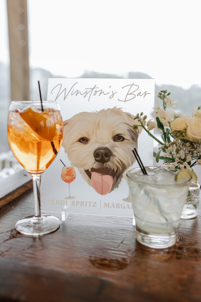 Custom wedding bar sign featuring a dog illustration, signature cocktails, and floral arrangements on a wooden bar.