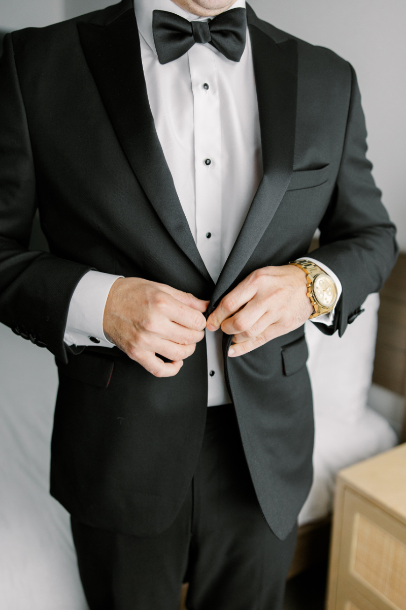 Groom adjusting black tuxedo jacket with bow tie and gold watch.