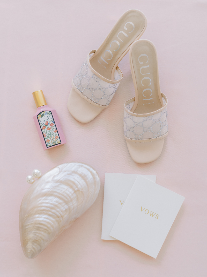 Bridal details with white heels, perfume, vow books, and shell clutch.