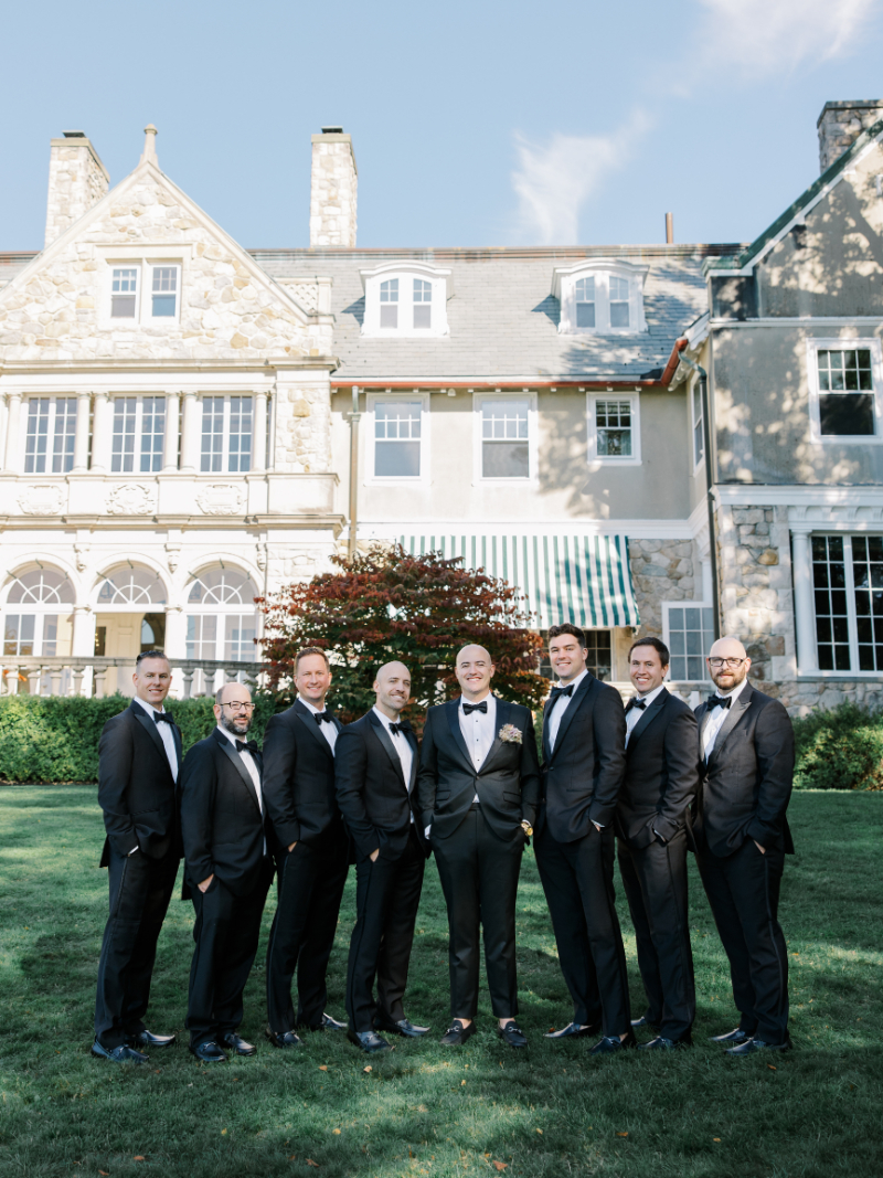 Groom and groomsmen in black tuxedos standing on lawn outside mansion.