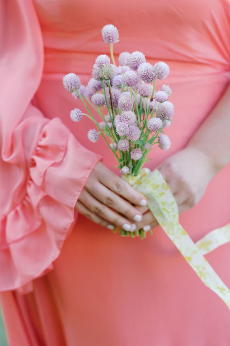 Bridesmaid in coral dress holding small lilac bouquet with ribbon.