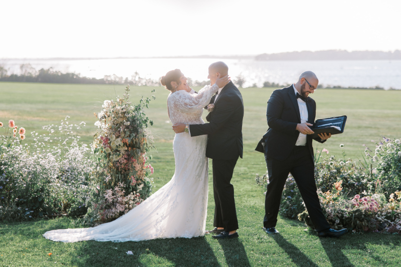 Bride and groom embracing during outdoor ceremony with floral arch backdrop.
