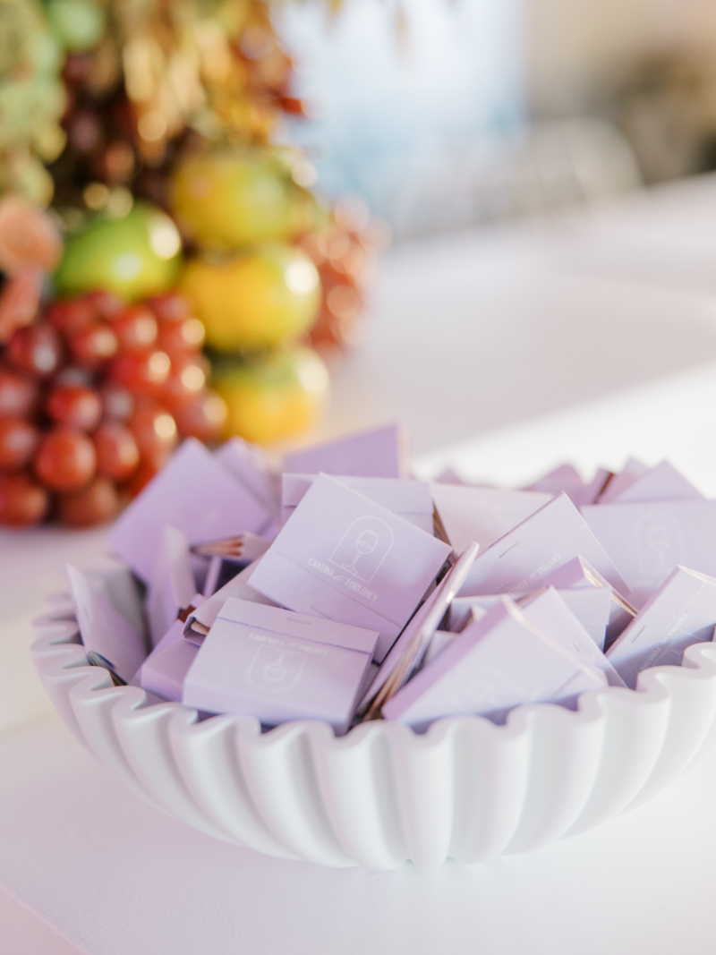 Bowl of lavender matchbooks displayed beside colorful fruit and floral arrangement.