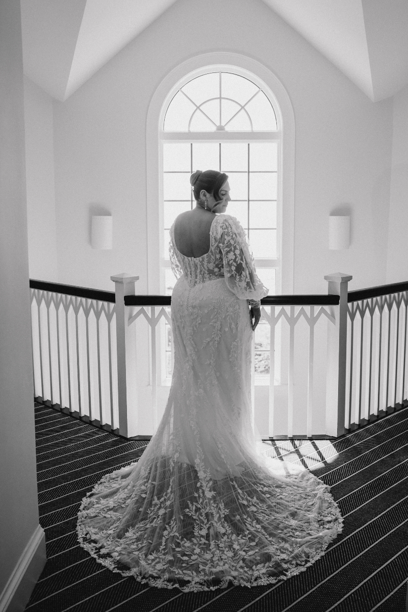 Bride in lace gown standing by arched window with dramatic train.