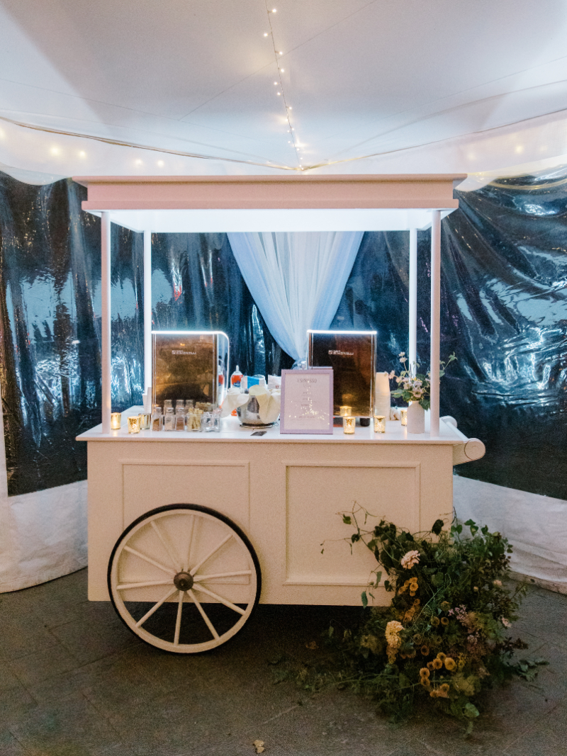 White espresso cart setup with candles and florals inside reception tent.