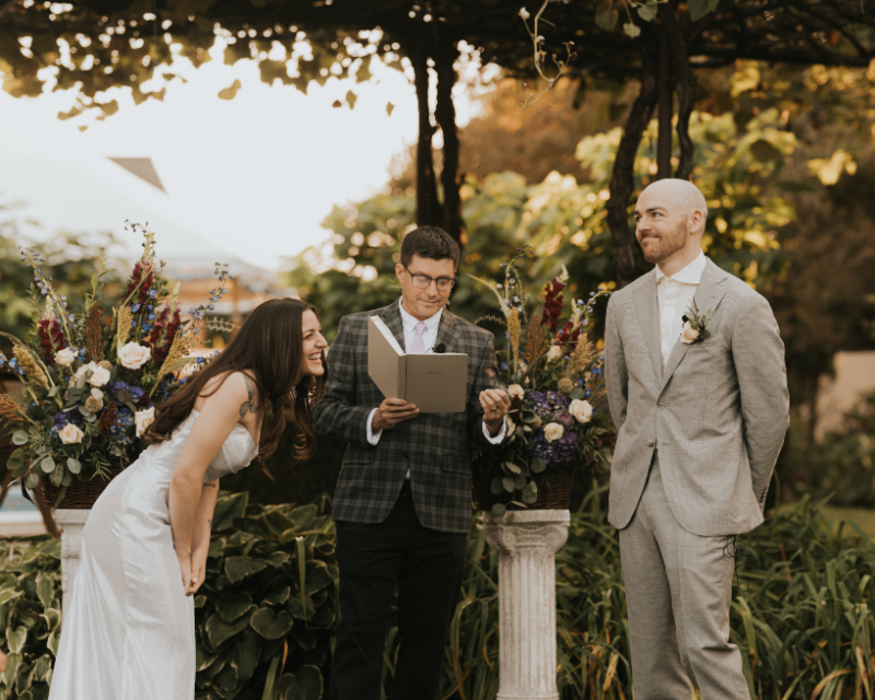 Officiant reading vows during Italian countryside wedding under grape vine canopy.
