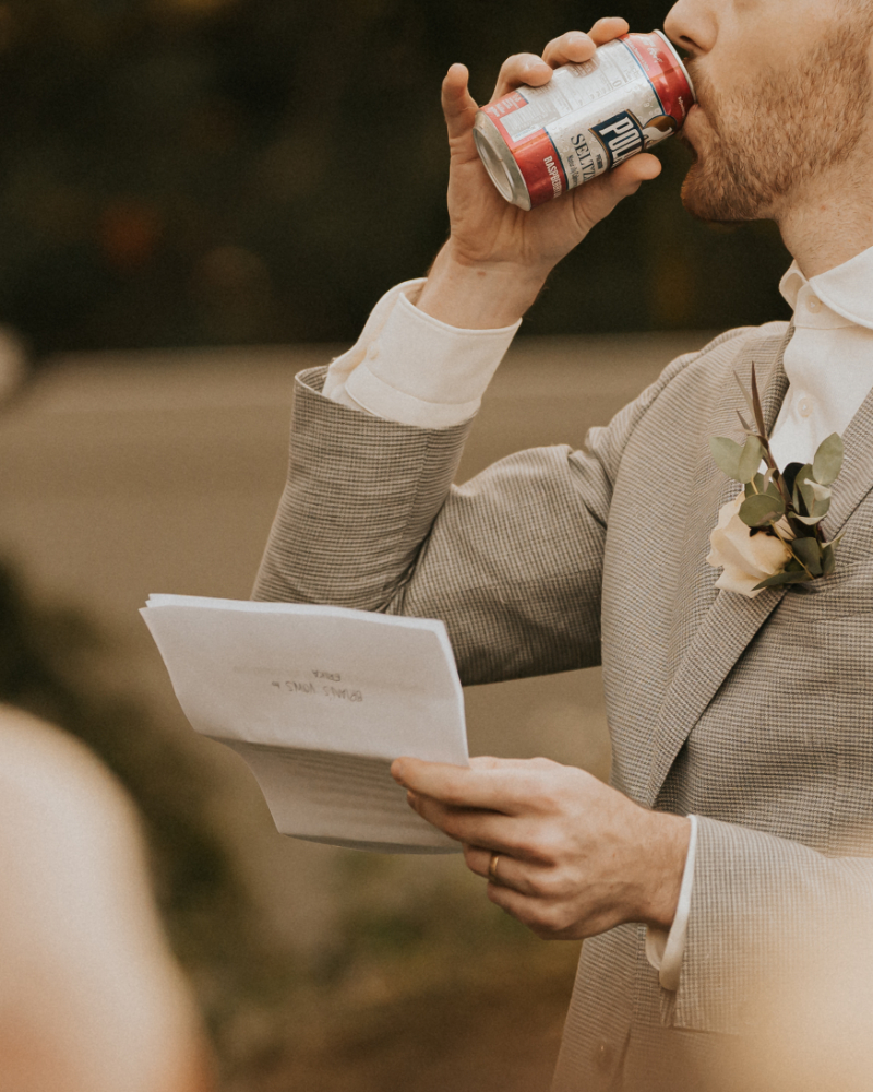 Groom reading vows and sipping drink during Italian countryside wedding ceremony.