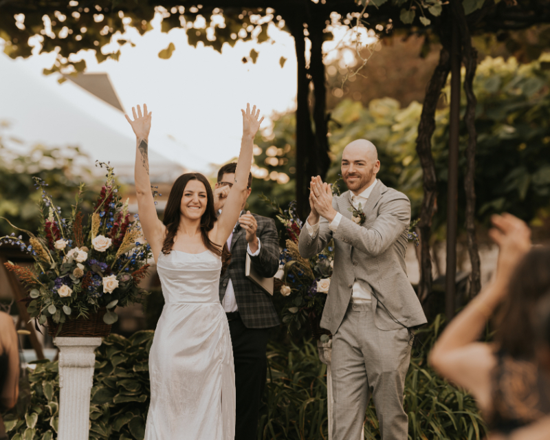 Bride celebrating with raised arms as groom applauds at Italian countryside wedding ceremony.