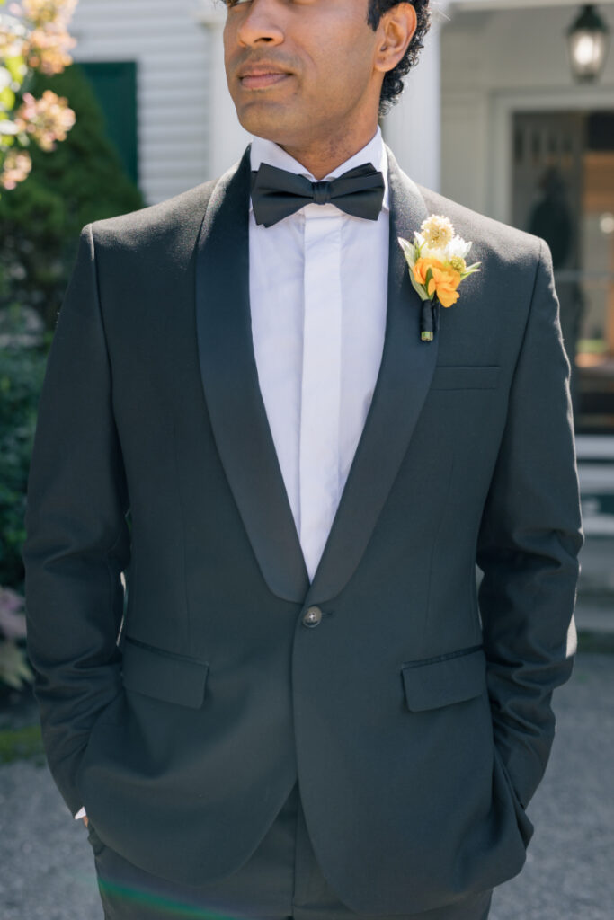 Groom in black tuxedo with a yellow boutonnière, standing with hands in pockets before the ceremony.
