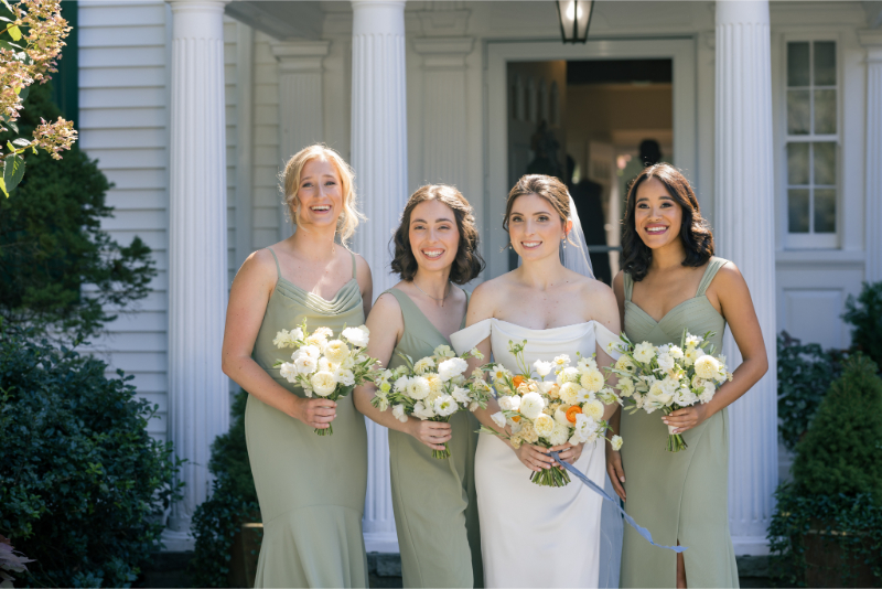 Bride standing with three bridesmaids in sage green dresses, all holding white and yellow bouquets.